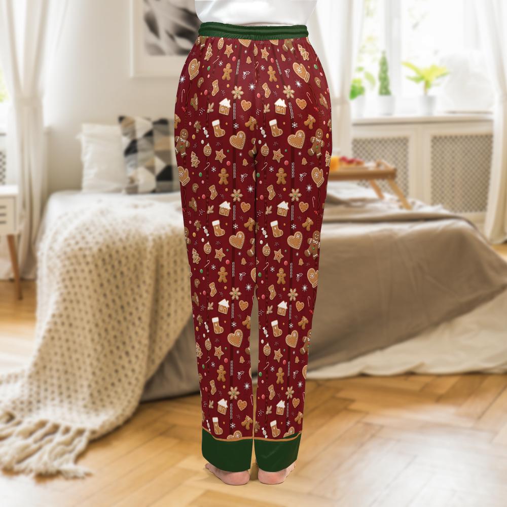 A person shown from the back wearing burgundy holiday pajama pants with a green waistband and cuffs, covered in a gingerbread-themed print of cookies, hearts, and boxes, standing in a bedroom.