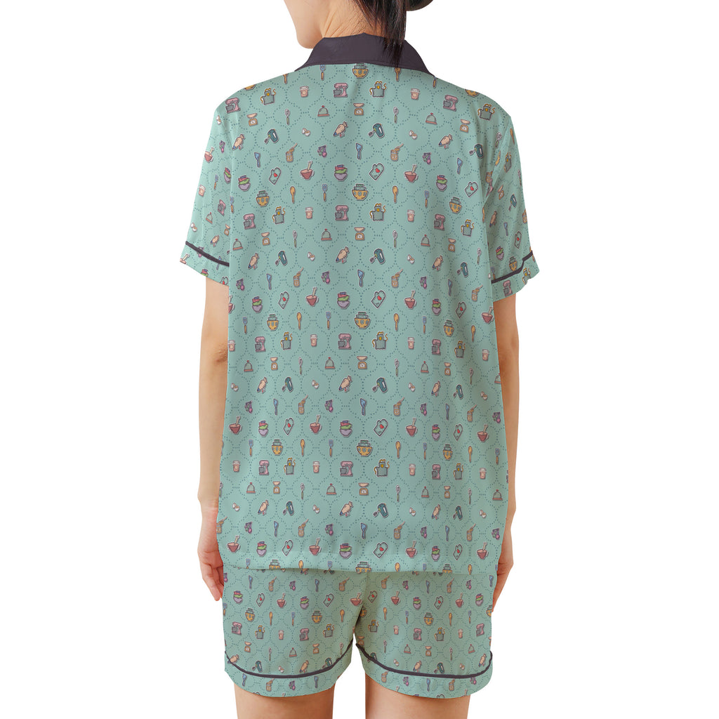 A woman shown from the back wearing a matching two-piece pajama set. The set consists of a short-sleeve top and shorts in a light sage green color with a dense, small multicolor pattern of vintage-style illustrations, featuring a dark charcoal collar and piping.