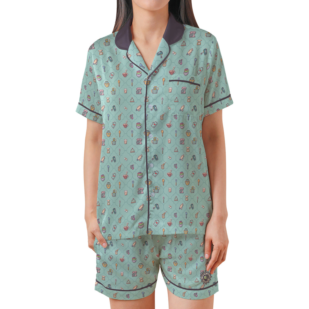 A woman modeling a two-piece pajama set featuring a mint green short-sleeve top and matching shorts with a small multicolor pattern, dark purple collar, and dark purple piping.