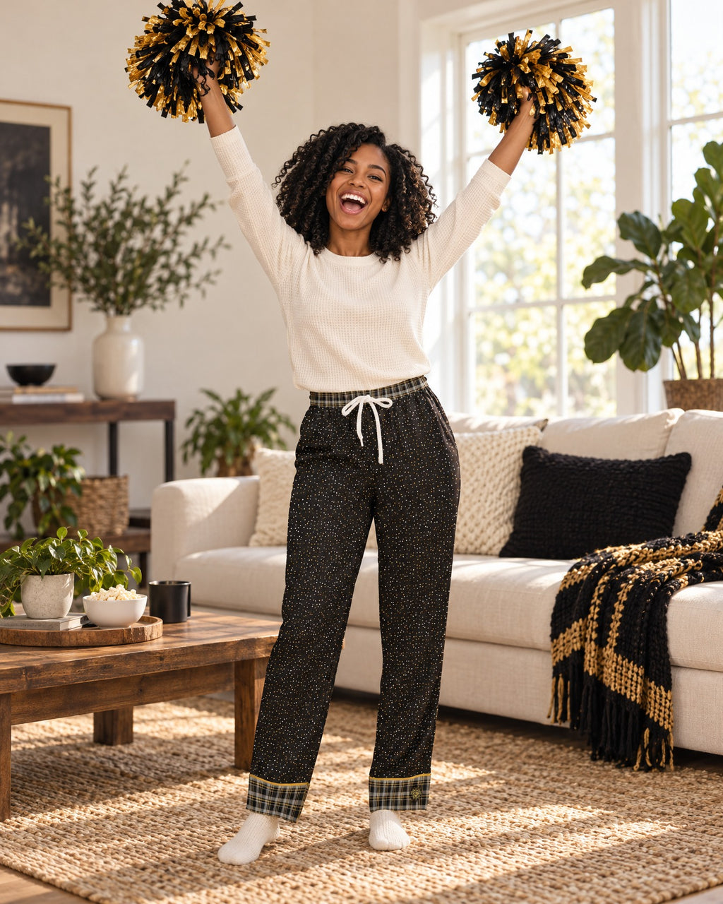 Woman cheering with both arms raised and plain pom-poms in the Black & Butterscotch satin pajama pants and a cream knit top