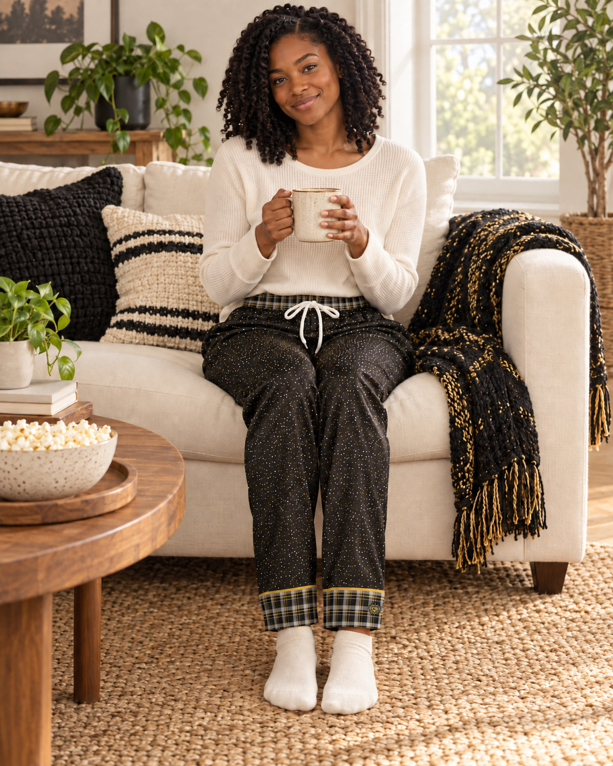 Woman sitting on a sofa holding a mug, wearing the Black & Butterscotch satin pajama pants with a cream knit top and showing the plaid cuffs