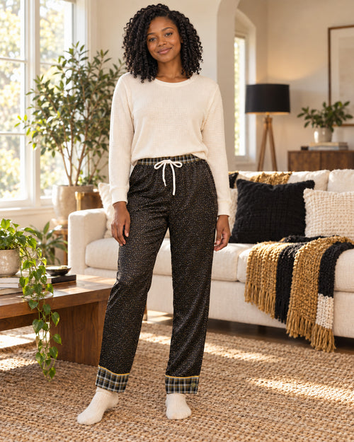 Woman standing in a sunlit living room wearing the Black & Butterscotch satin pajama pants with a cream knit top