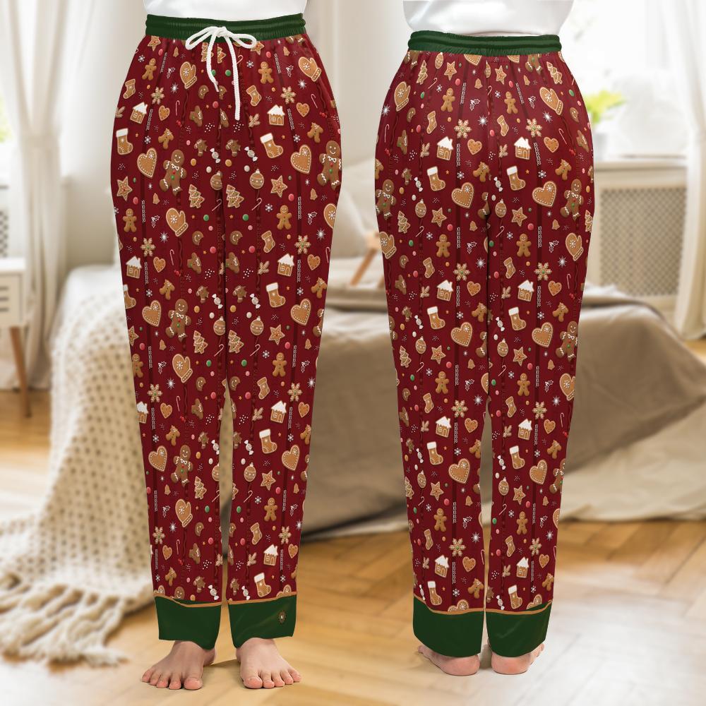 A pair of Christmas-themed pajama pants shown from both the front and back views, worn by a model in a cozy bedroom setting. The pants are burgundy with a festive pattern of gingerbread cookies, hearts, snowflakes, and small houses, featuring a dark green waistband and cuffs with a white drawstring.
