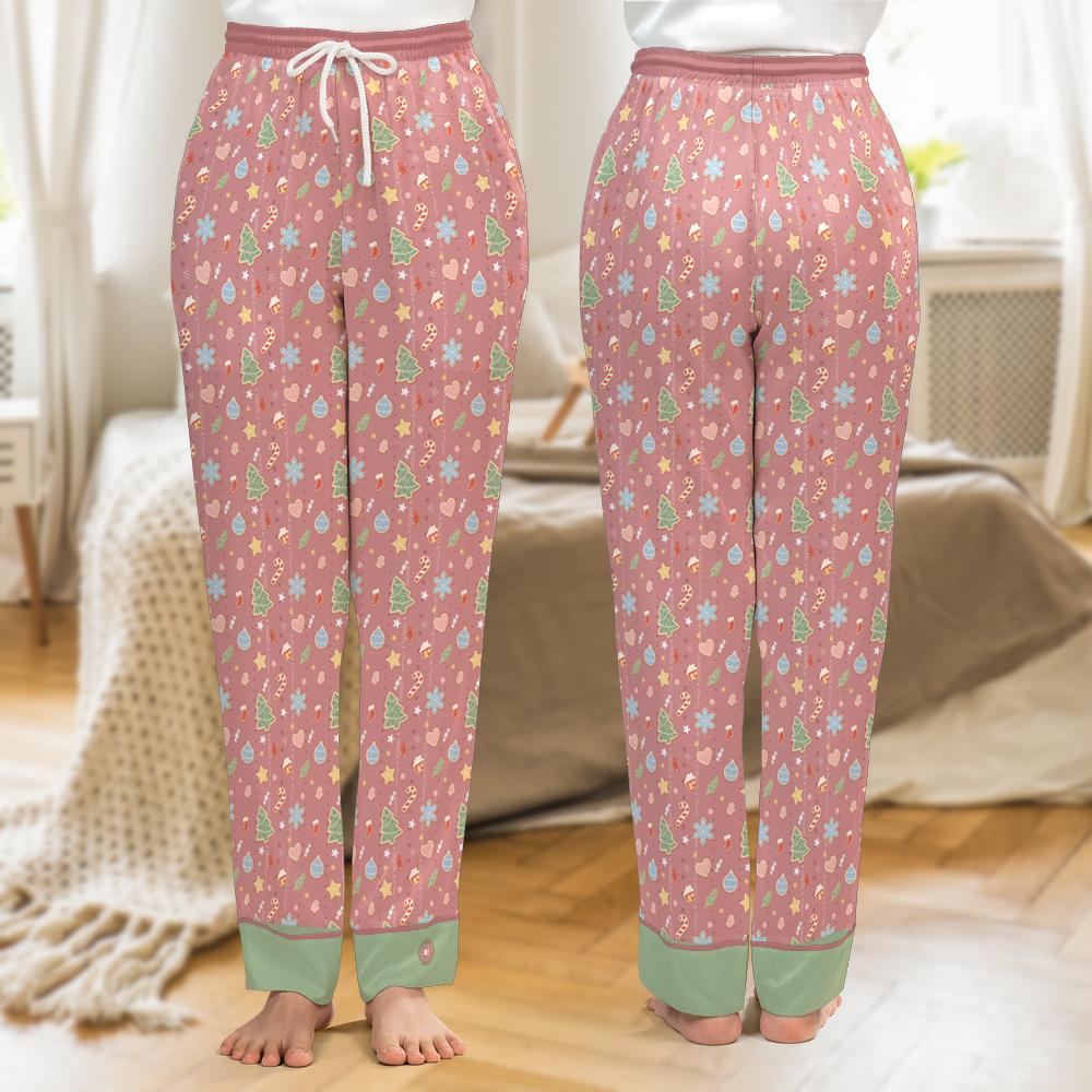 A pair of pink patterned pajama pants shown from the front and back, worn by a model standing barefoot in a bedroom setting. The pants feature a festive holiday print with pine trees, ornaments, and snowflakes in green, blue, yellow, and white, with sage green cuffs and a white drawstring.
