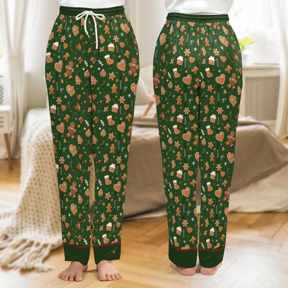 A pair of green holiday-themed pajama pants with a gingerbread print, shown from the waist down on a model in a bedroom setting. The pants feature a white drawstring waist, a festive pattern of gingerbread men, cookies, hearts, and snowflakes, and ribbed cuffs.