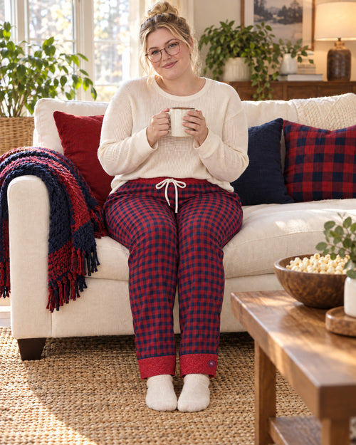 Woman sitting on a sofa holding a mug, wearing the Cherry & Navy satin pajama pants with a cream knit top and showing the plaid cuffs