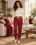 Woman standing in a sunlit living room wearing the Crimson Cocoa satin pajama pants with a cream knit top