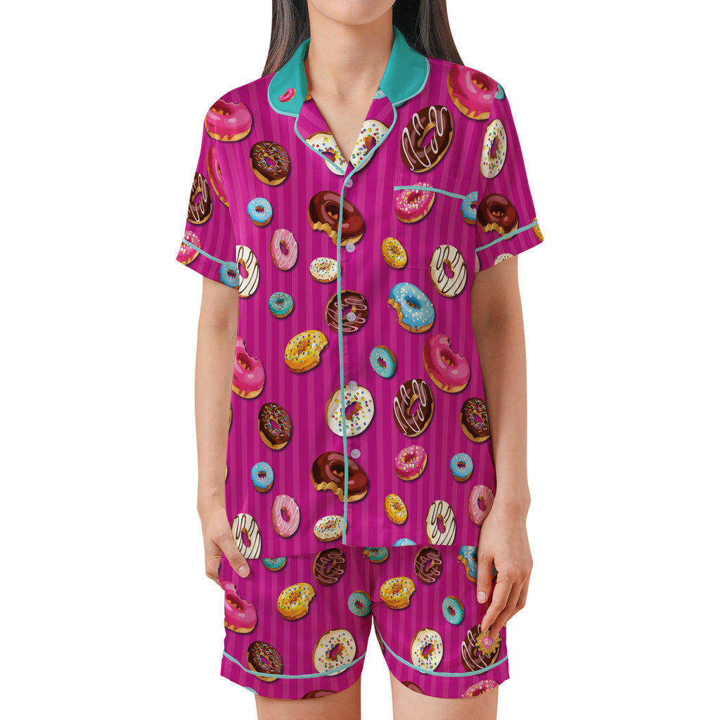 A woman wearing a matching short-sleeve pajama top and shorts set with a bright pink and purple striped background covered in a colorful donut print, featuring teal trim and collar.