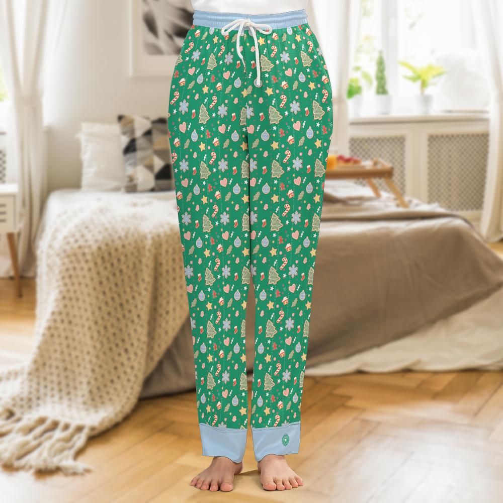 A person standing barefoot in a bedroom wearing green pajama pants with a repeating holiday tree and ornament print, a light blue drawstring waist, and light blue cuffs.