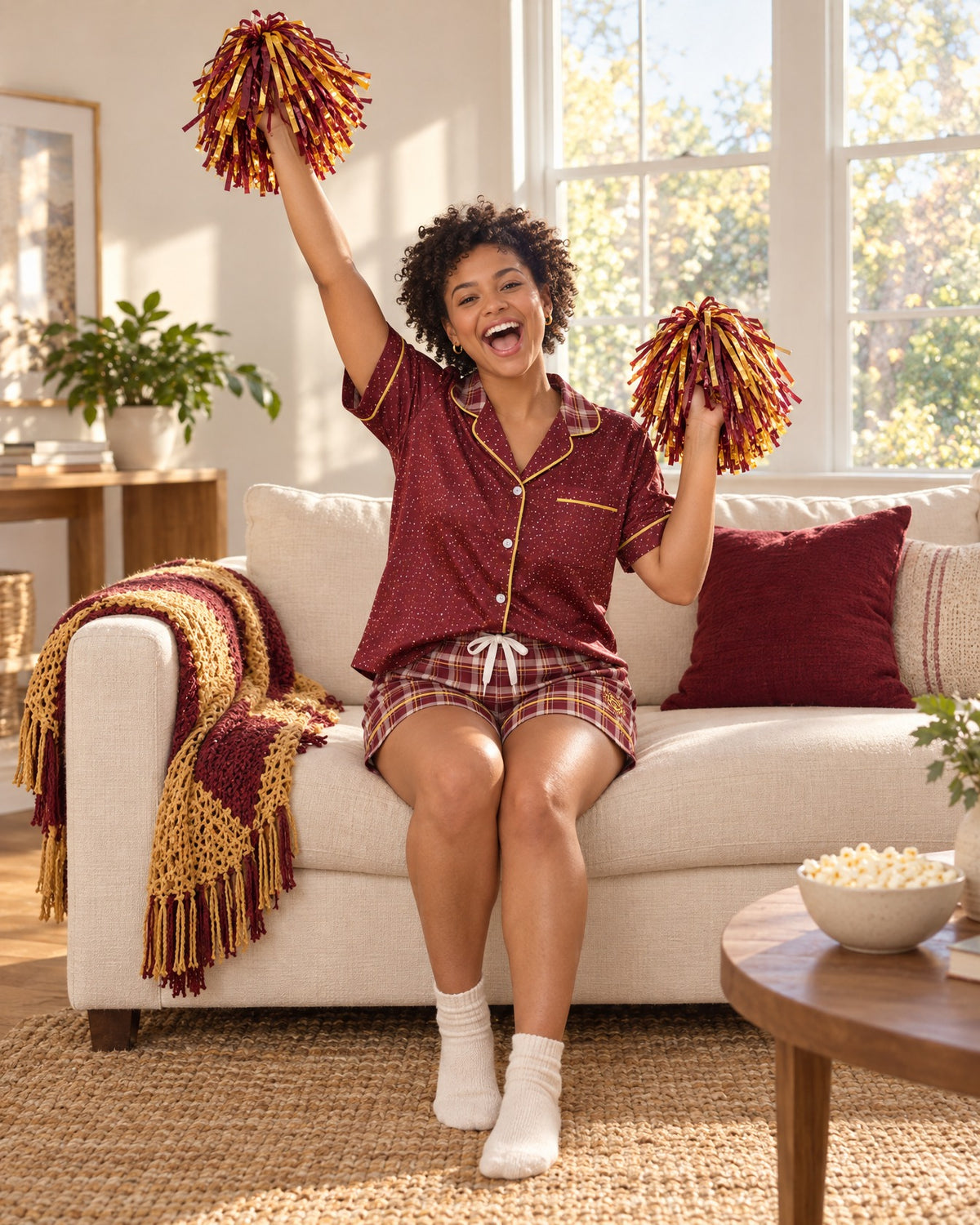Woman cheering on a sofa in the Garnet & Honey satin pajama set holding team-color pom-poms