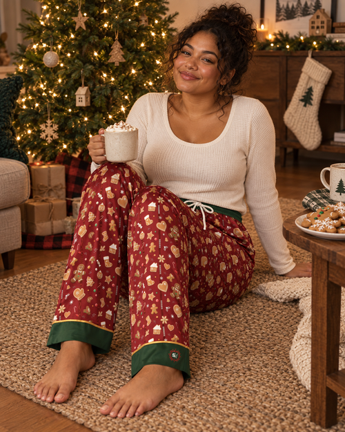A woman sits on a rug in a cozy living room wearing a holiday-themed pajama set. The set includes a cream ribbed long-sleeve top and red pajama pants with a gold festive pattern and green cuffs.