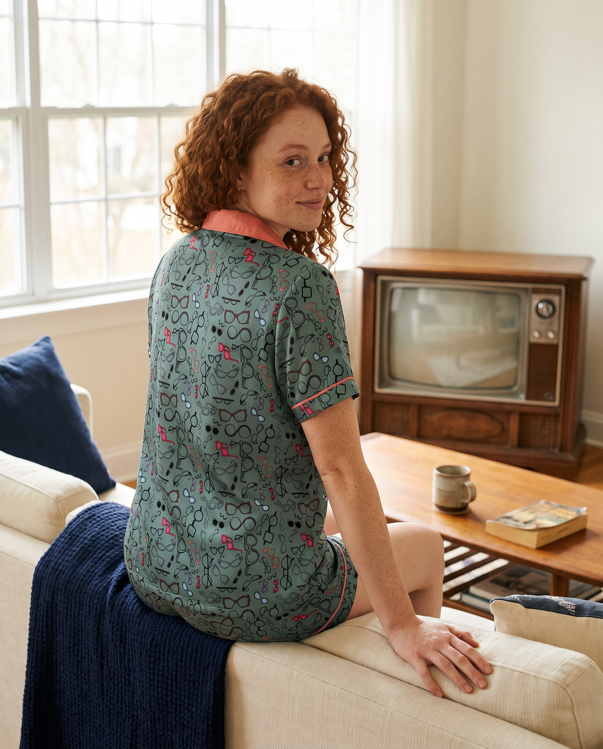 A woman with curly red hair sits on a cream sofa in a living room, wearing a short-sleeve pajama top with a green base and a pink, red, and blue abstract pattern. She is turned to the side, looking over her shoulder.