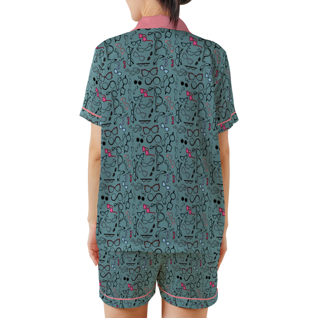 A model shown from the back wearing a matching two-piece pajama set. The set consists of a short-sleeve top and shorts in a dark teal-green color, decorated with a dense pattern of black cartoon faces, glasses, and bows, accented with pink trim on the collar, sleeve cuffs, and shorts hem.