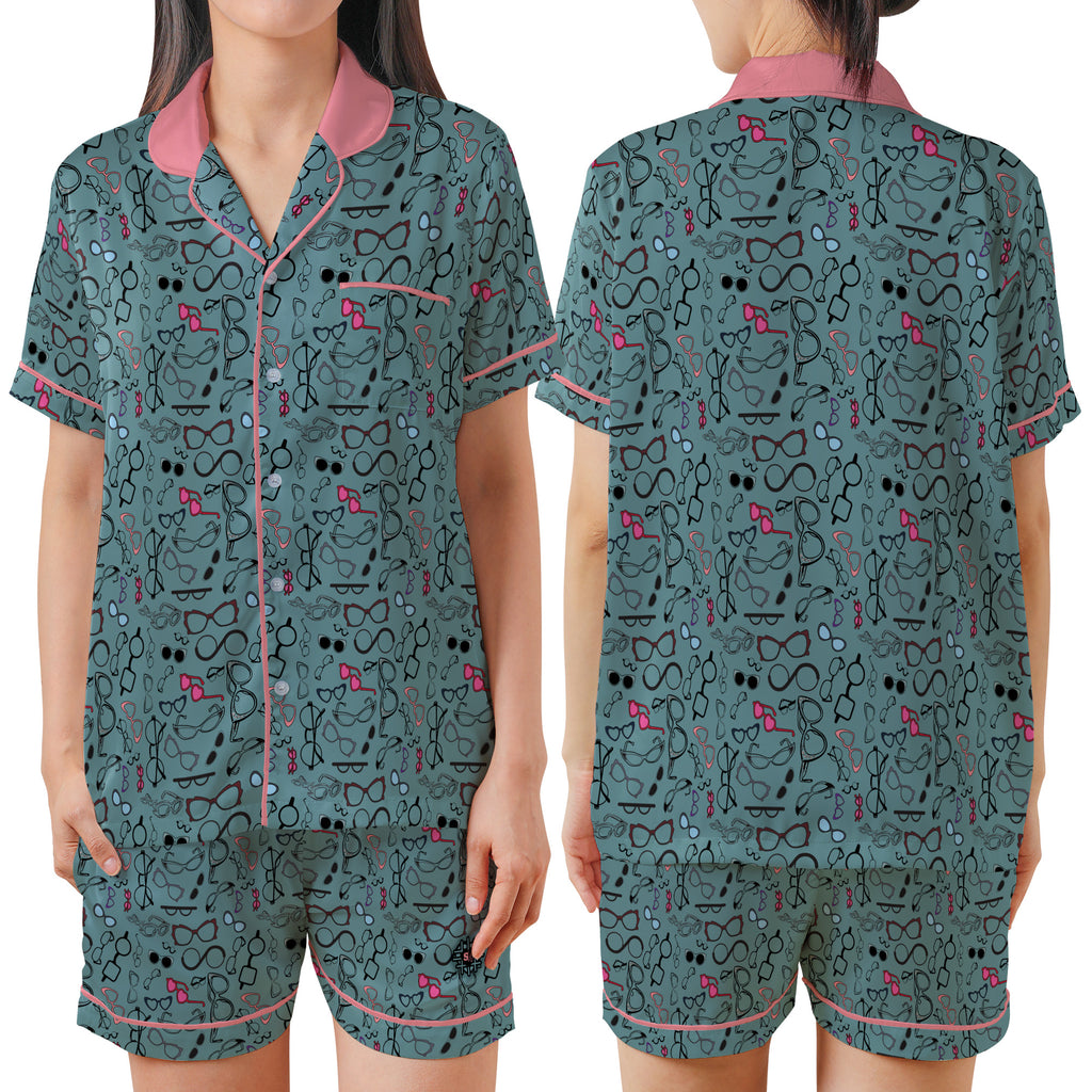 A women's pajama set shown on a model from the front and back, featuring a dark teal-green background with a black cartoon cat pattern and pink accents on the collar, trim, and small details.