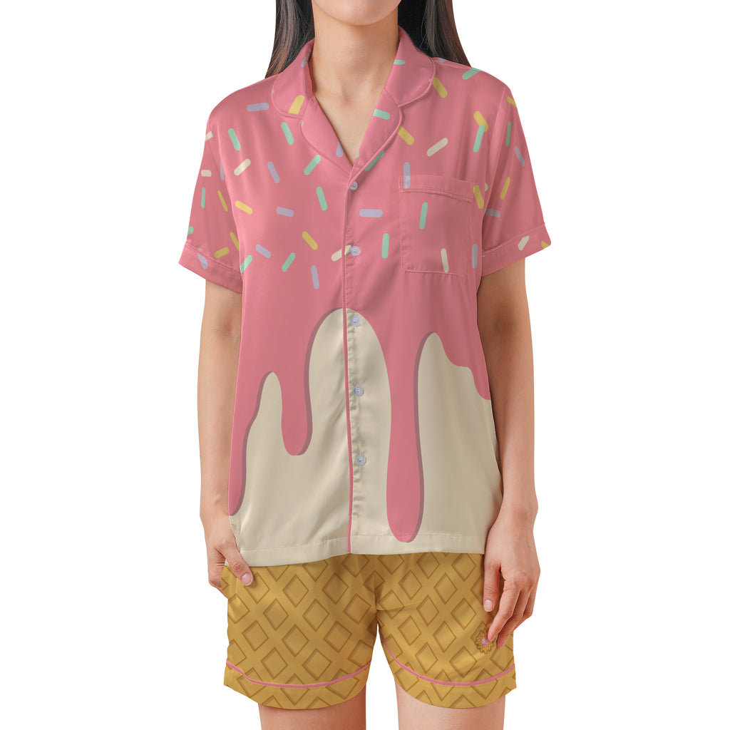 A woman modeling a short-sleeve pajama shirt with a pink and cream ice cream drip design and multicolored sprinkle pattern, paired with yellow patterned shorts against a plain white background.