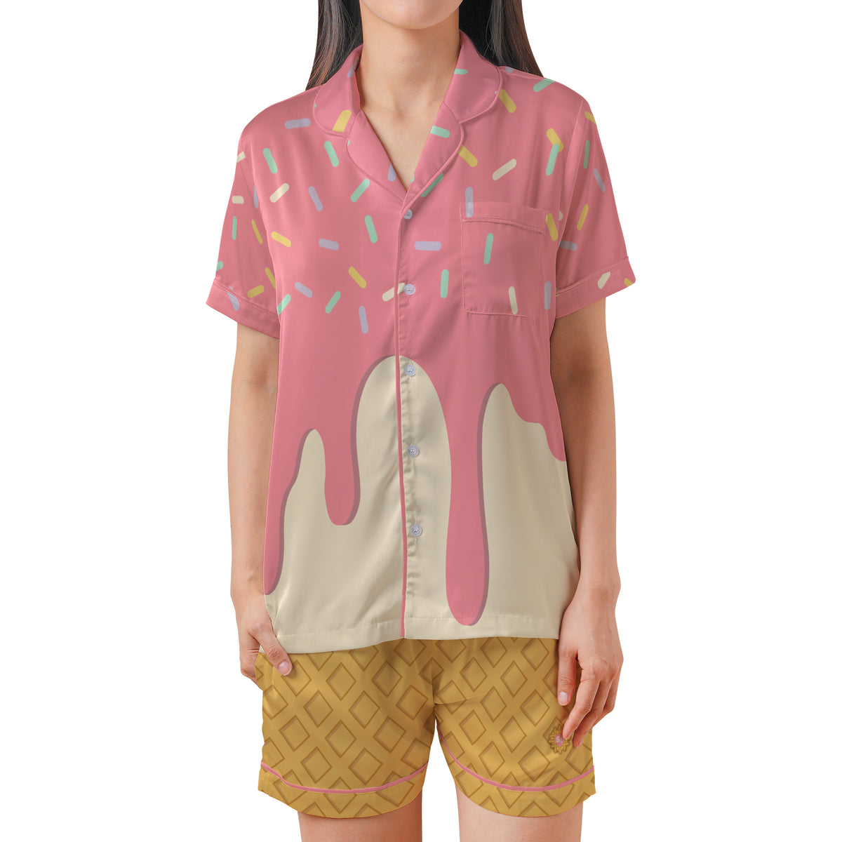 A woman modeling a short-sleeve pajama shirt with a pink and cream ice cream drip design and multicolored sprinkle pattern, paired with yellow patterned shorts against a plain white background.