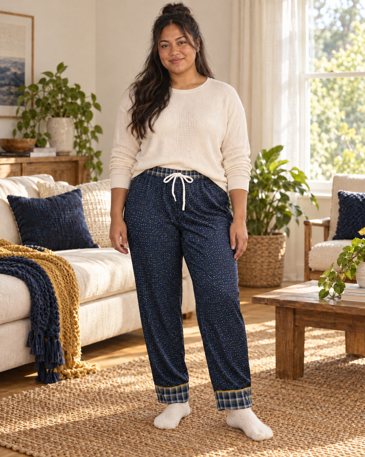 Woman standing in a sunlit living room wearing the Navy & Brass satin pajama pants with a cream knit top