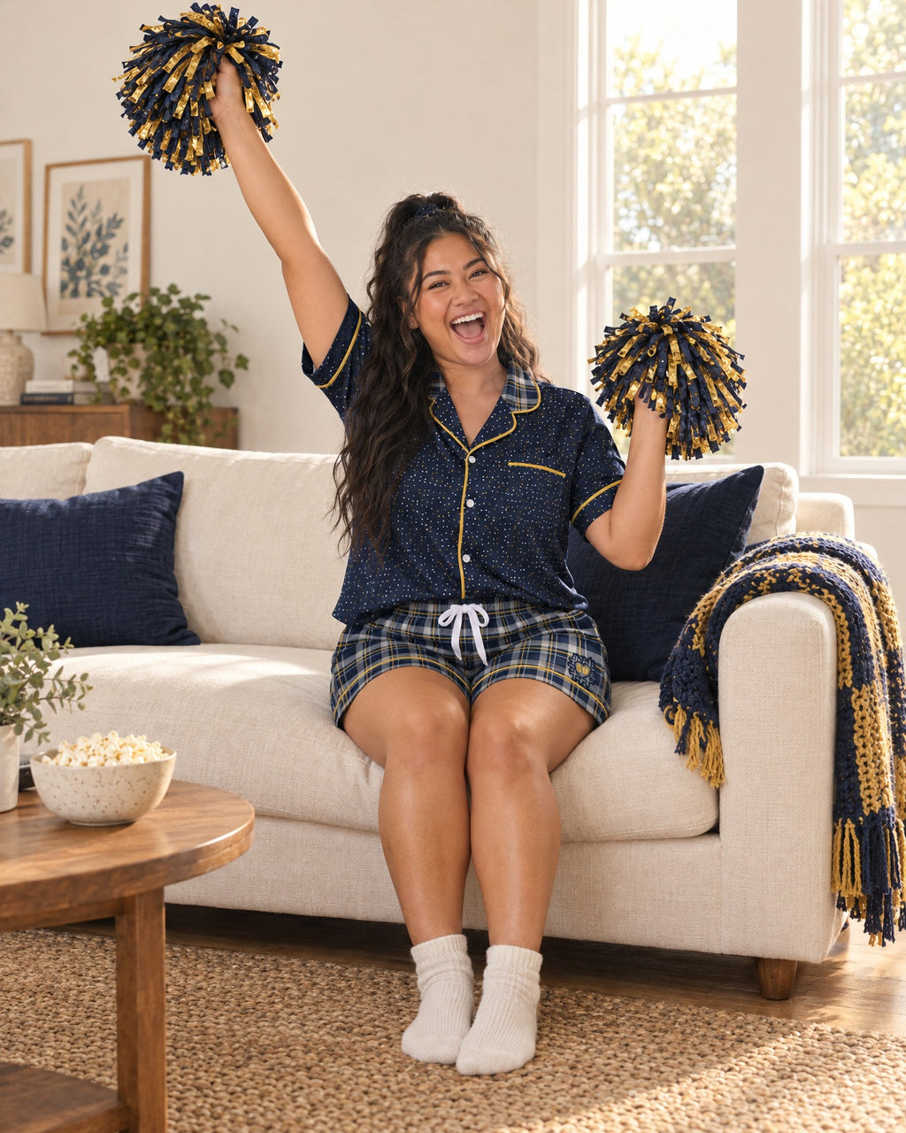 Woman cheering on a sofa in the Navy & Brass satin pajama set holding team-color pom-poms