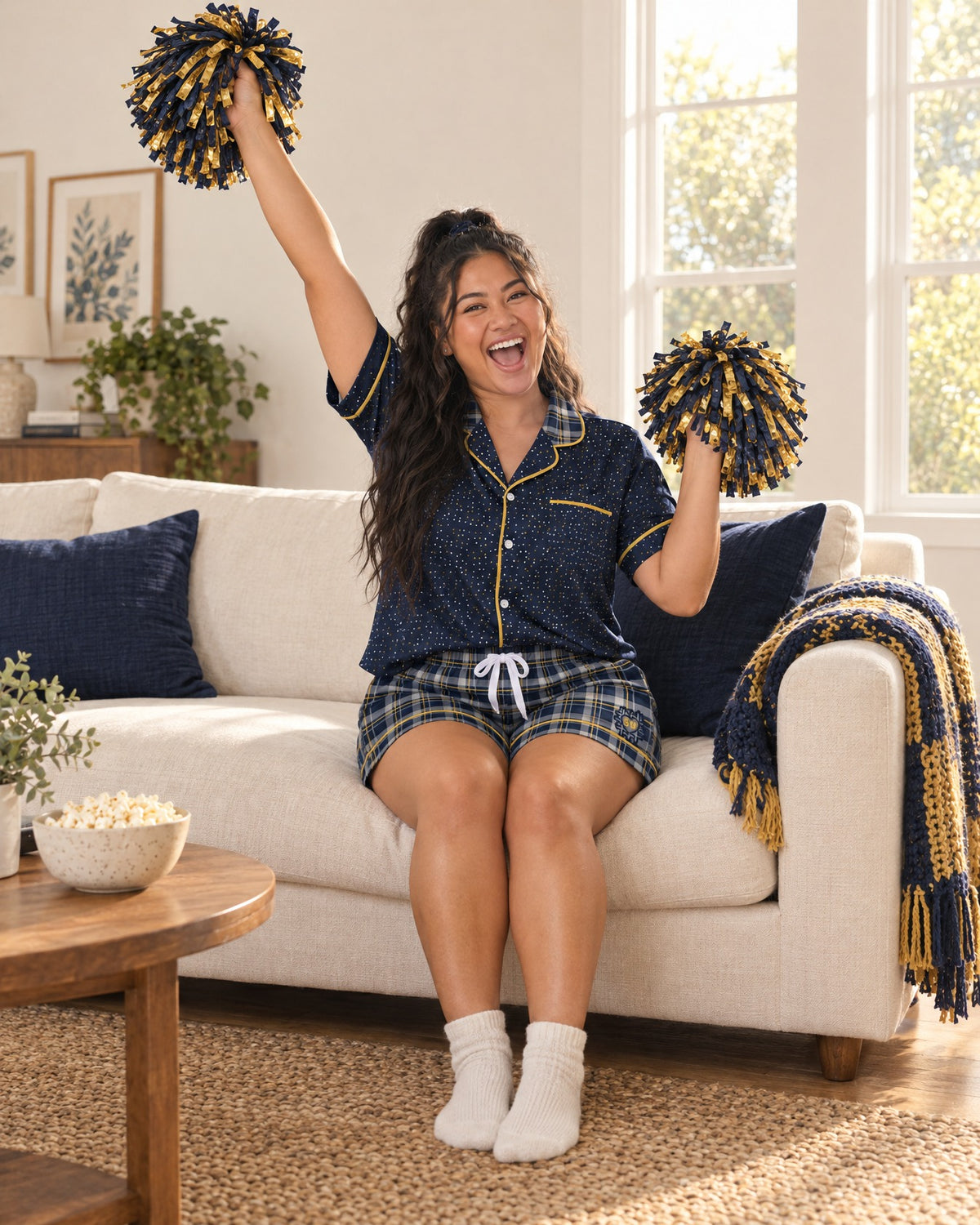 Woman cheering on a sofa in the Navy & Brass satin pajama set holding team-color pom-poms