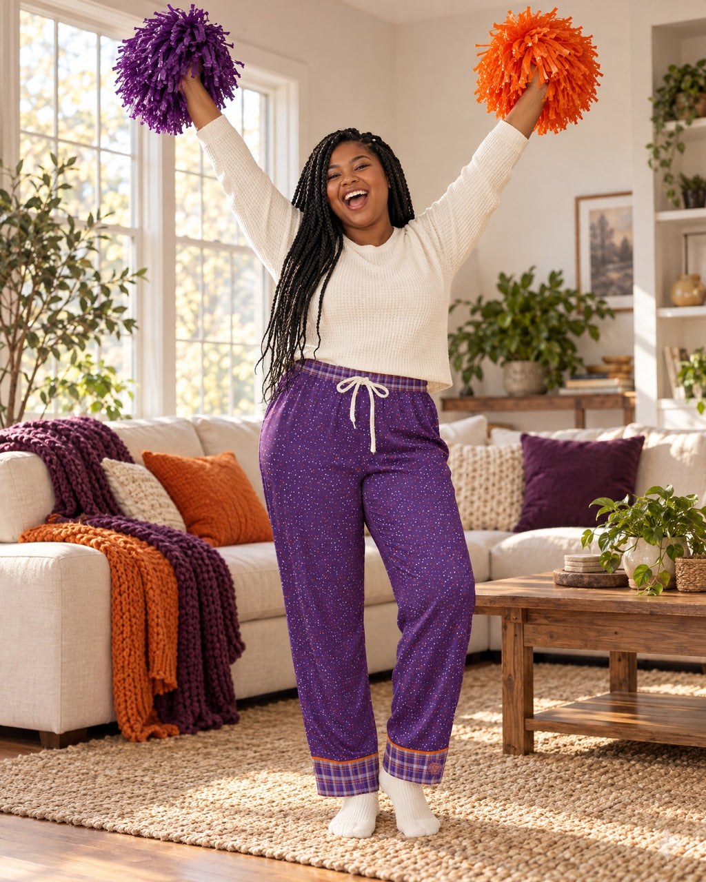 Woman cheering with both arms raised and plain pom-poms in the Orange & Plum satin pajama pants and a cream knit top