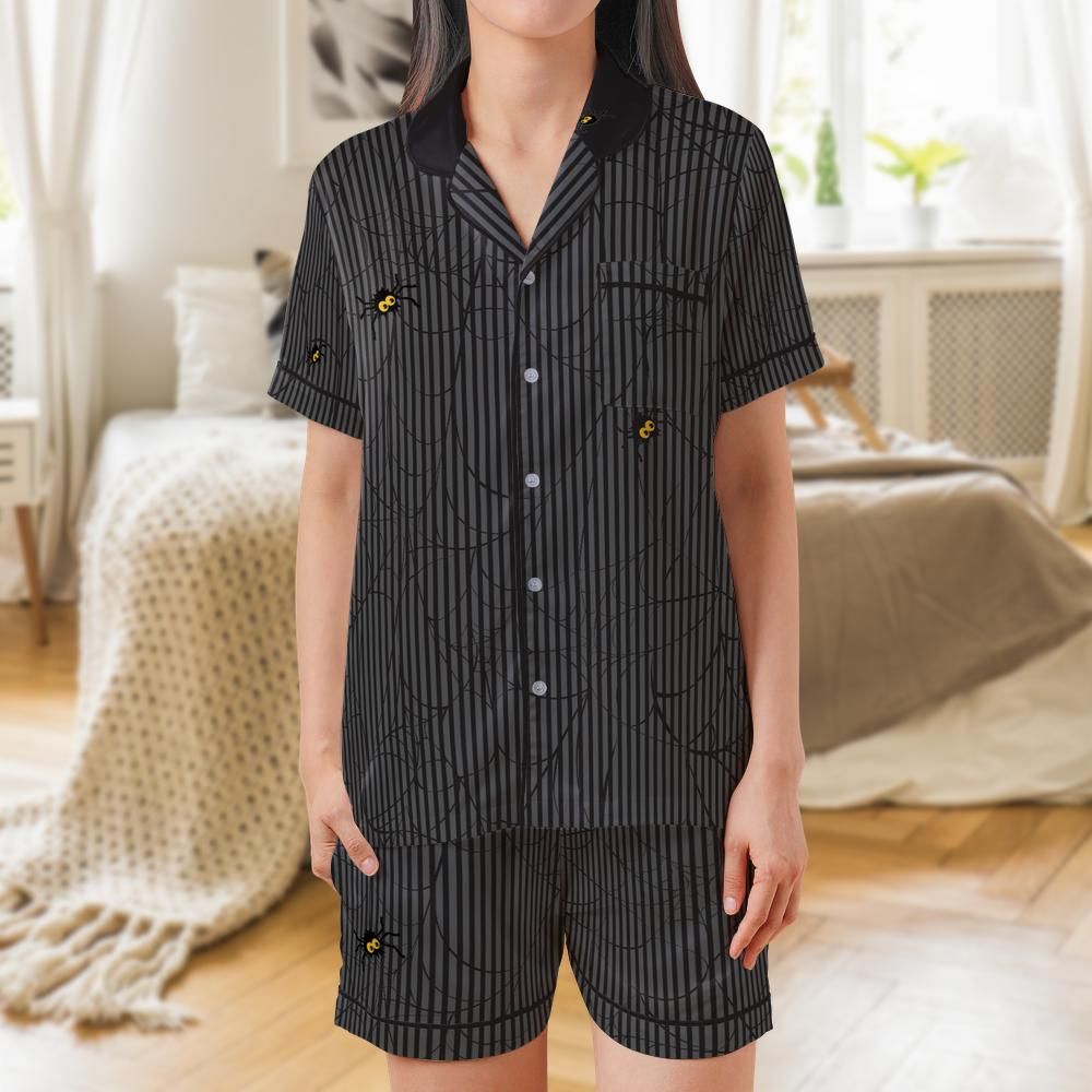 A woman wearing a matching short-sleeve button-front pajama shirt and shorts set with a black and gray vertical stripe spider pattern, shown in a softly lit bedroom setting.