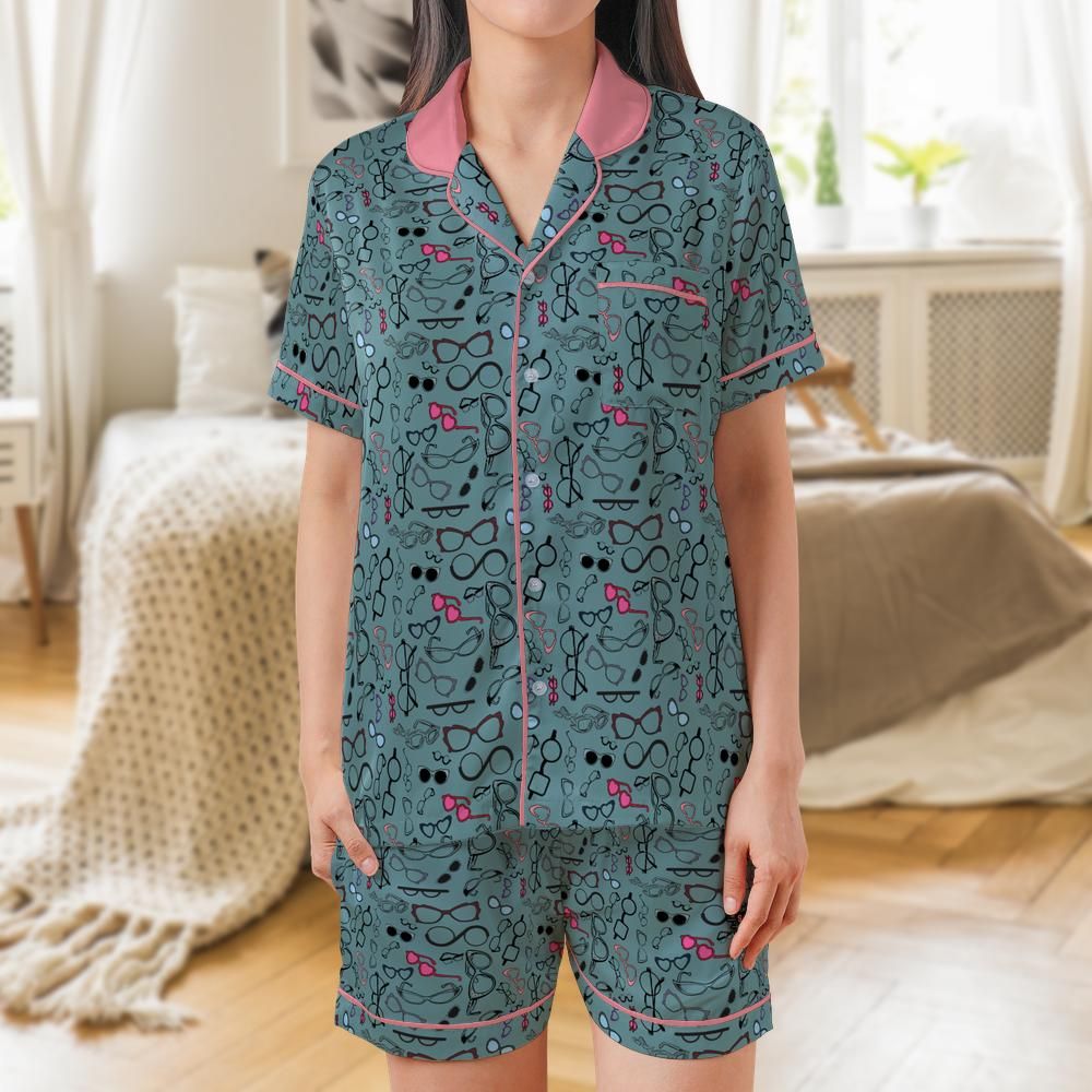 A woman wearing a matching short-sleeve pajama set with a teal background, pink collar and piping, and a black-and-pink abstract pattern, standing in a softly lit bedroom.