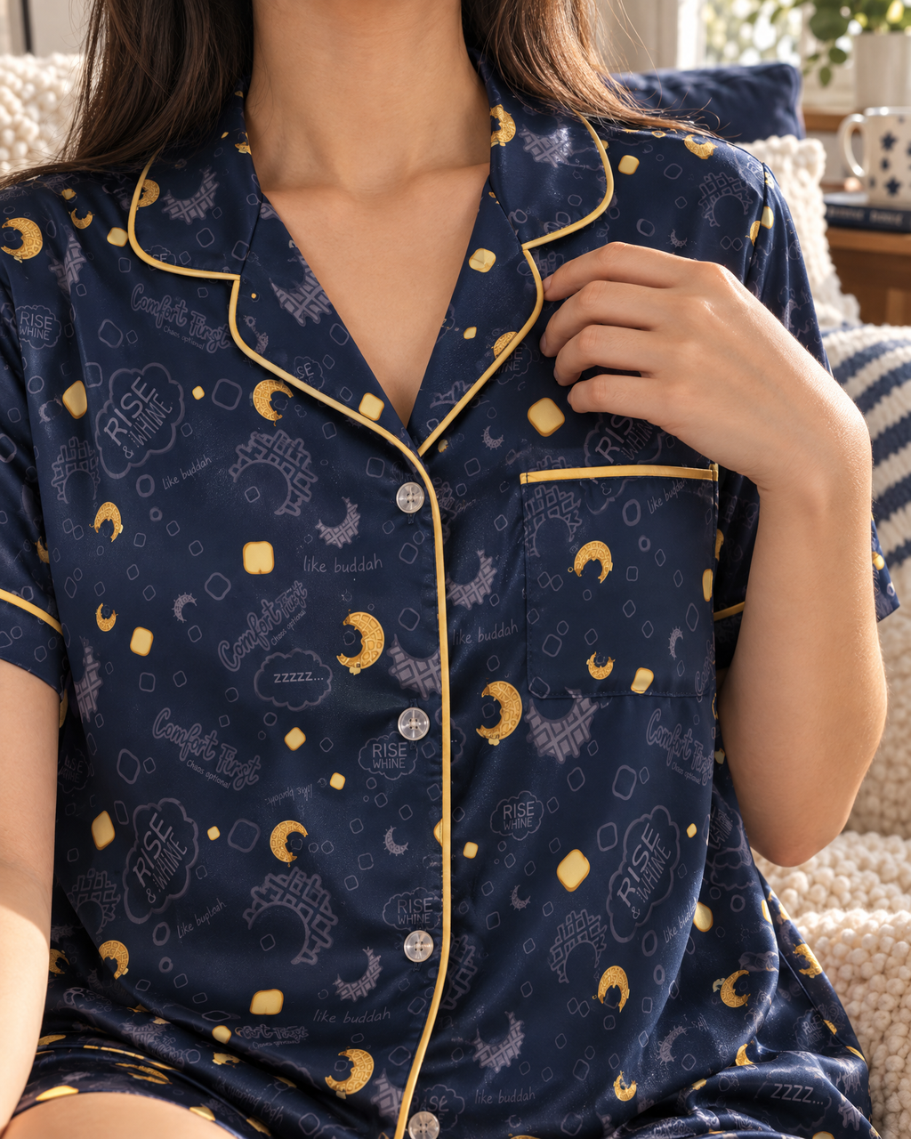 A woman wearing a navy blue short-sleeve pajama shirt with a gold moon waffles and butter pats pattern, sitting on a cream sofa in a cozy living room with an old television in the background.
