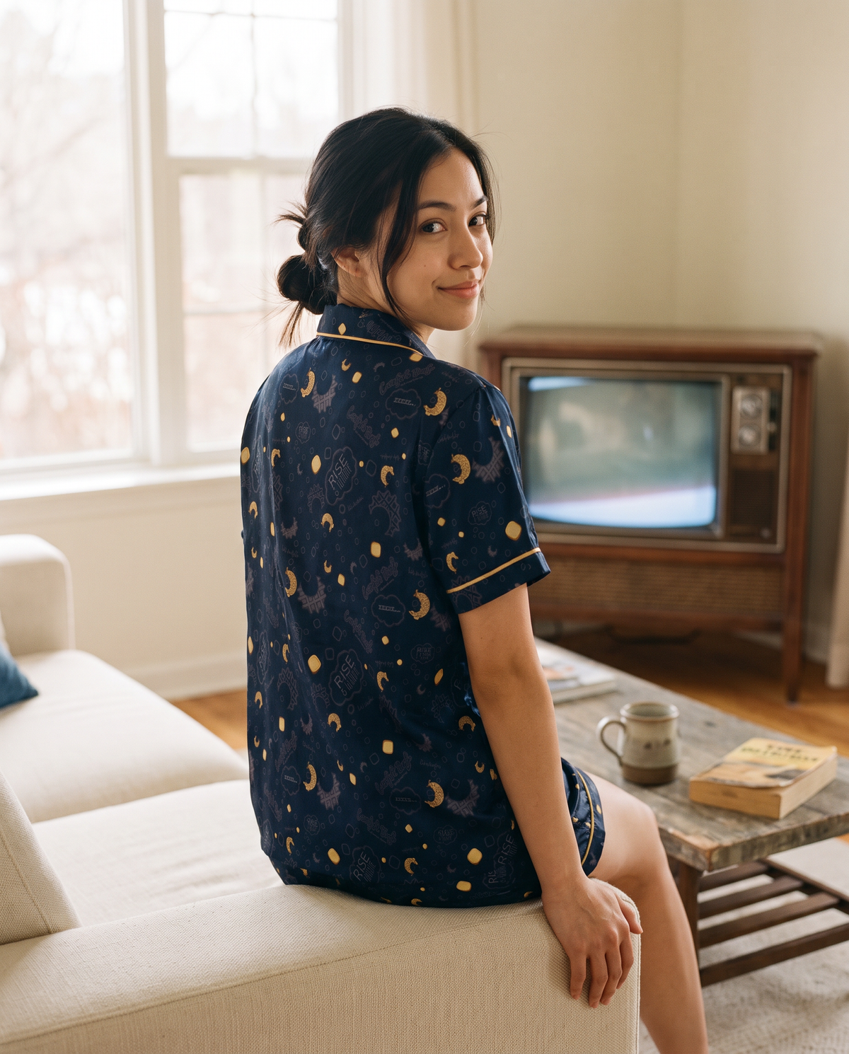 A woman wearing a navy blue short-sleeve pajama shirt with a gold moon waffles and butter pats pattern, sitting on a cream sofa in a cozy living room with an old television in the background.