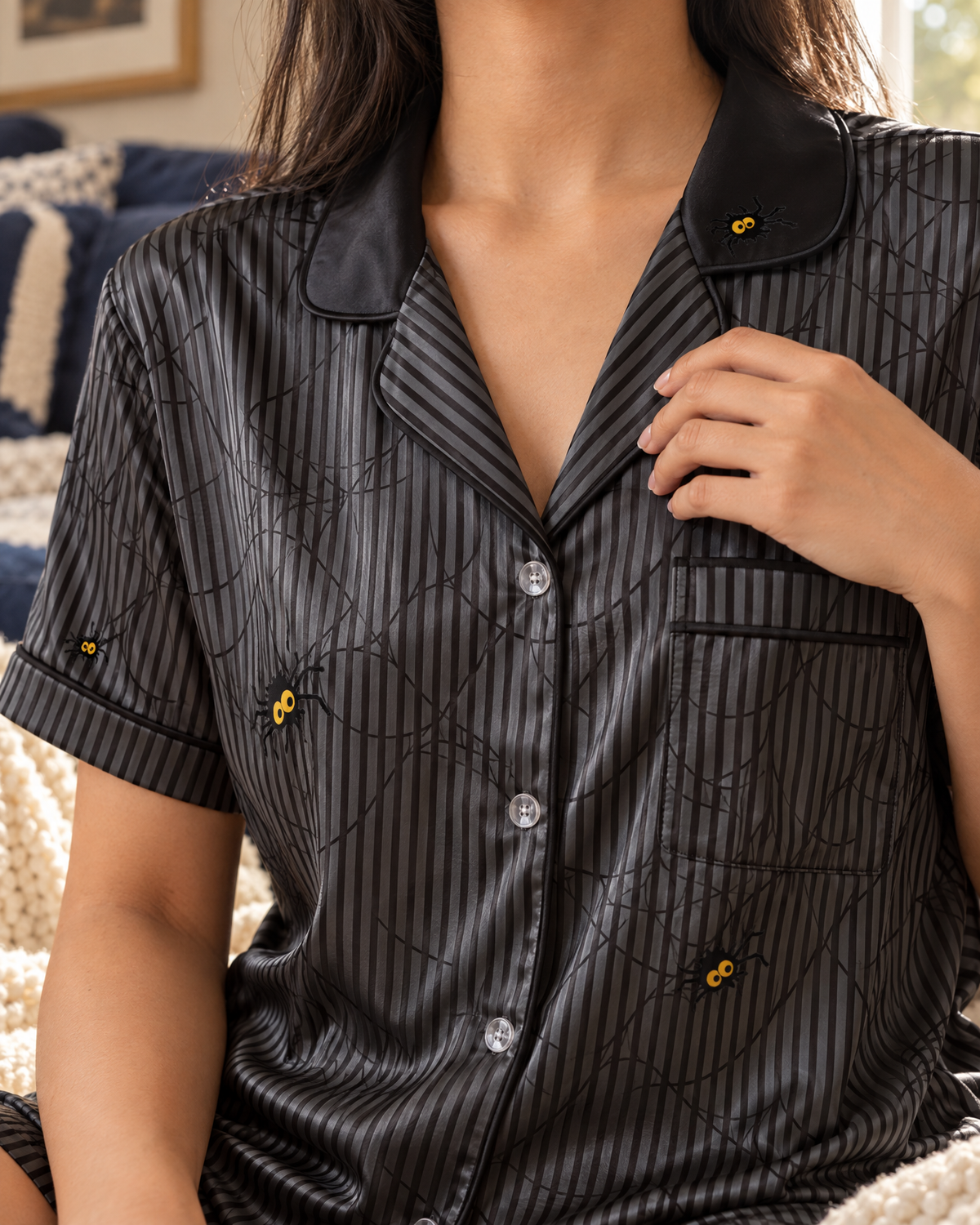 A close-up of a person wearing a black short-sleeve pajama shirt with thin vertical stripes, a black collar, and small embroidered yellow spiders on the chest and sleeves. The person is seated indoors with a bed and pillows visible in the background.