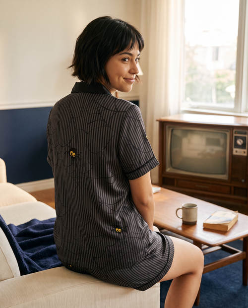 A woman wearing a short-sleeve black and gray striped shirt with yellow buttons, sitting on a beige sofa in a retro-living room with a vintage television and wooden coffee table.
