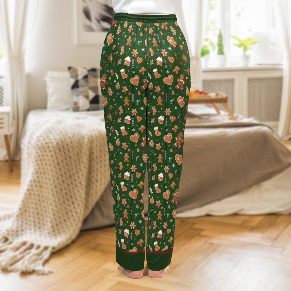 A person shown from the back wearing dark green holiday pajama pants with a repeating print of gingerbread men, cookies, hearts, and small houses, standing in a softly lit bedroom.